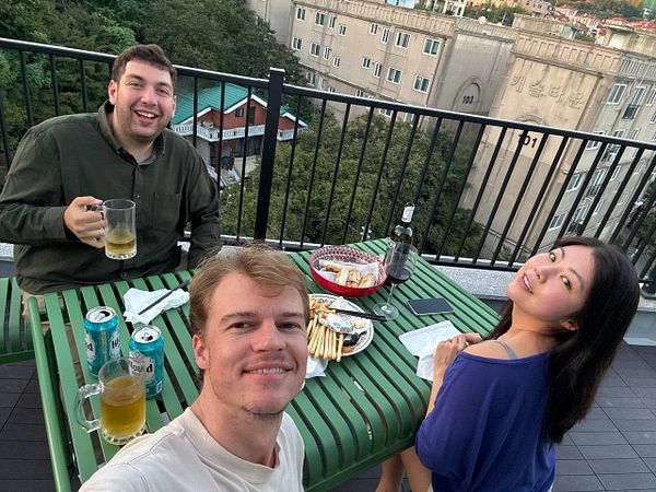 Mini home balcony setup with an air fryer cooking food, a paper plate with fried snacks, and a glass of orange drink on a wooden outdoor table.