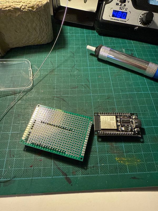 The image depicts a workspace for soldering, featuring a circuit board and a microcontroller.