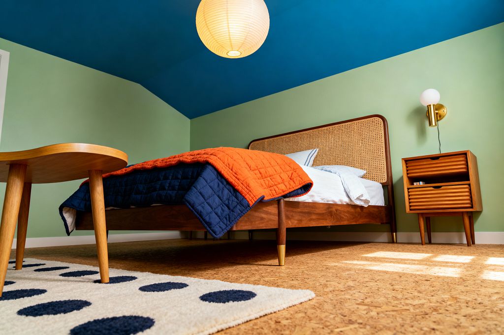 A mid-century modern kids’ room features a walnut bed with a woven cane headboard, white sheets, and a reversible orange and navy quilt. Light sage walls contrast a deep blue ceiling and a round paper lantern, while a brass globe sconce hangs over a fluted walnut nightstand; cork flooring and a cream rug with navy polka dots sit under a small wood stool and sunlight from an unseen window.