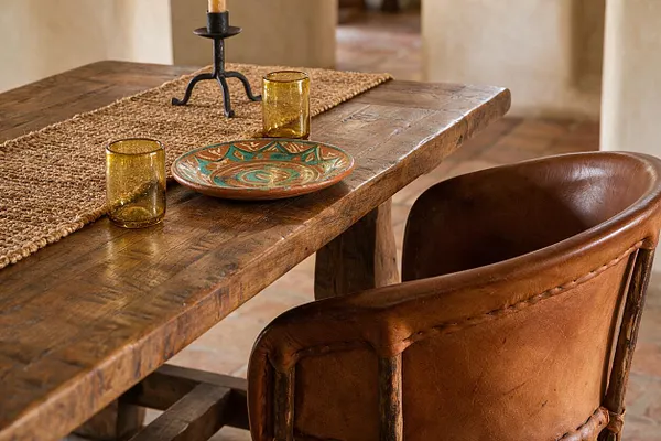 A rustic Southwestern dining vignette features a thick reclaimed wood table with a woven jute runner, amber glass tumblers, a painted ceramic plate, and a black metal candlestick. A brown leather chair sits in the foreground against warm neutral walls and terracotta-toned flooring.