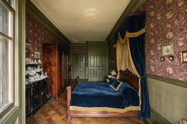 A narrow Victorian-style guest bedroom with a carved wooden bed dressed in deep blue velvet and gold fringe, framed by heavy blue and gold drapery. The room features patterned floral wallpaper, olive green trim, parquet wood flooring, a black painted cabinet with decorative dishes, and a tall wooden wardrobe.