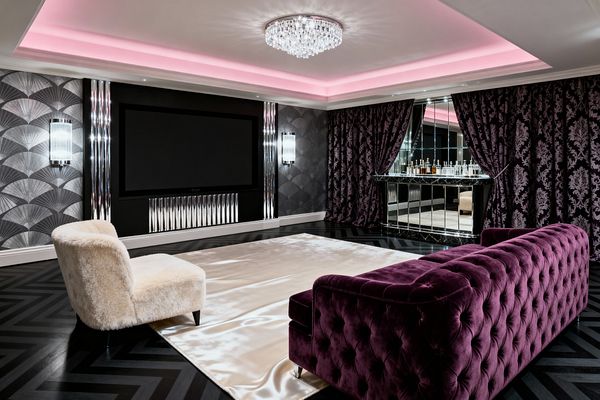 A glamorous media room pairs a deep plum tufted velvet sofa with an ivory faux‑fur lounge chair on a champagne satin rug over ebonized herringbone wood floors. A large black‑framed screen anchors an Art Deco wall with silver fan wallpaper, fluted chrome panels, and prismatic glass sconces, while a mirrored bar gleams behind purple damask curtains; a crystal ceiling light and pink cove lighting wash the tray ceiling, creating a dramatic vintage‑inspired atmosphere.