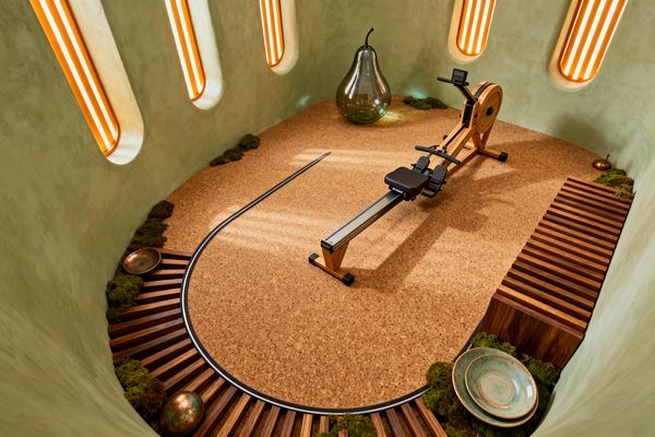 An oval, cave-like home gym features sage green plaster walls punctuated by tall rounded recesses that glow with vertical orange lighting, a cork floor traced by a curved metal channel, and a wood-and-metal rowing machine centered in the space. Moss-lined perimeter niches, striped hardwood benching, a large clear glass pear sculpture, and scattered copper and ceramic bowls create a dreamlike, organic atmosphere with warm, indirect light.