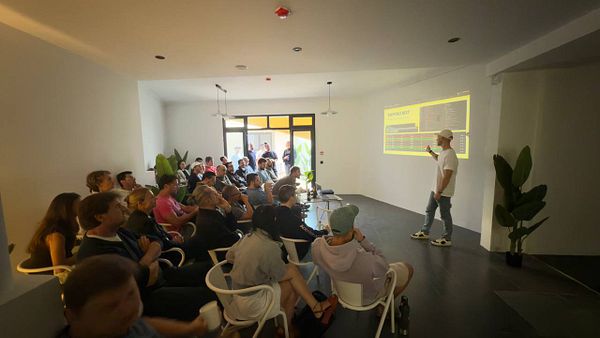 A tech meetup is taking place in a modern, well-lit room with a speaker presenting to an engaged audience.