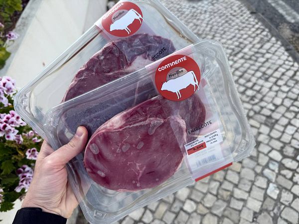 A person holds two packages of entrecôte steaks in a hand, surrounded by a floral background and cobblestone pavement.