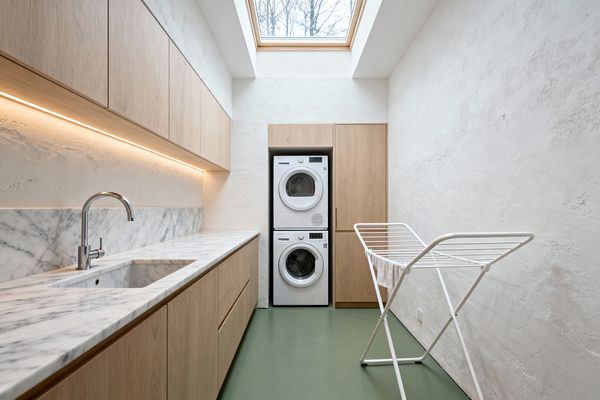 A narrow Scandinavian laundry room with light oak flat-panel cabinets, a white marble countertop and backsplash, and a chrome faucet over an undermount sink. Warm LED lighting runs beneath the upper cabinets. At the end, a white stacked washer and dryer are framed by tall oak storage. A large wood-framed skylight brings daylight onto textured white plaster walls and a matte sage-green floor. A white metal drying rack sits to the right.