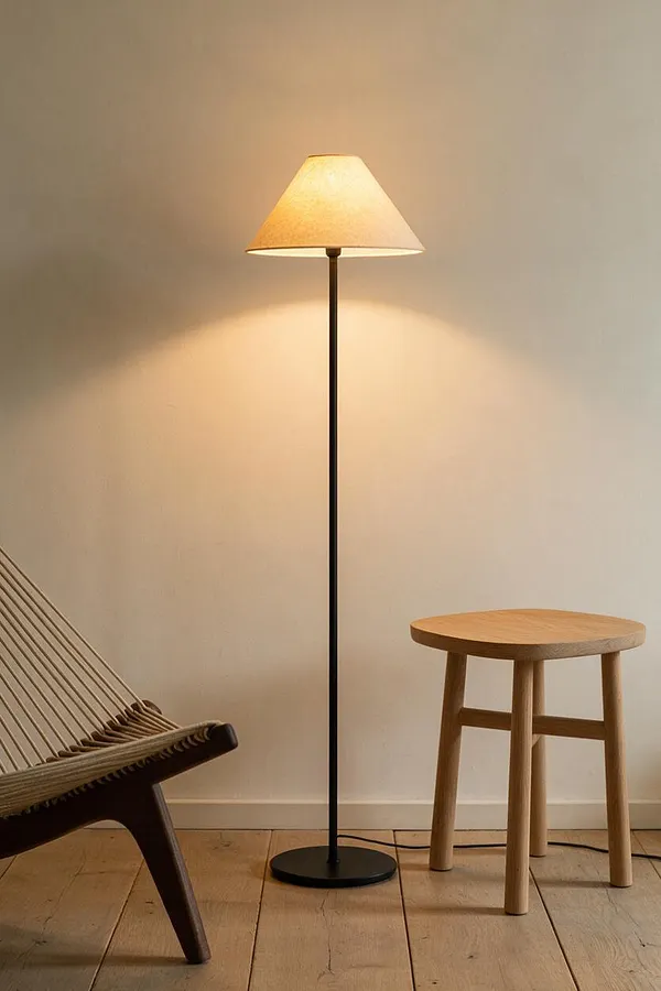 A minimalist Scandinavian corner with a tall black floor lamp topped by a soft beige shade, casting warm light on a plain white wall. A small natural wood stool sits beside it, and part of a woven lounge chair appears at the left edge over pale wood plank flooring.
