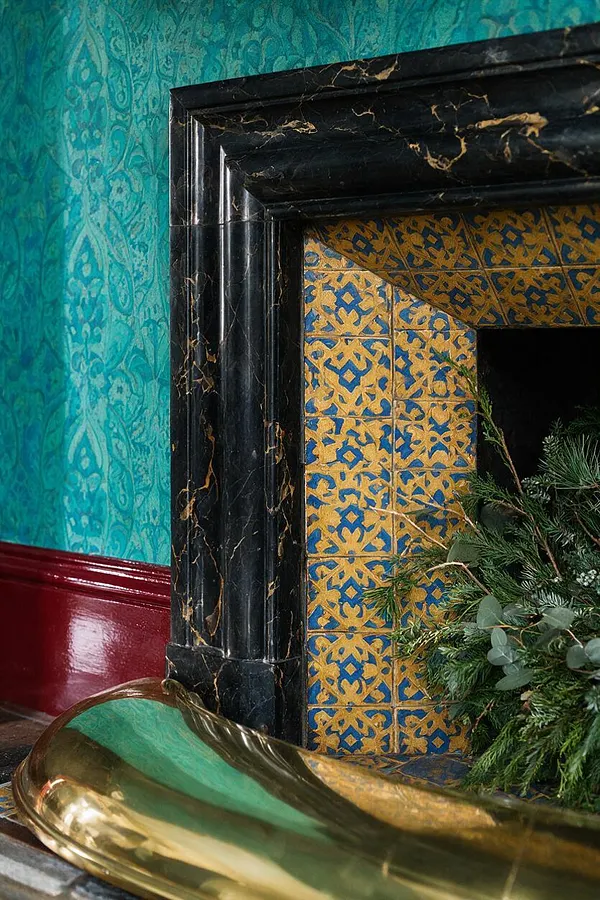 A richly layered interior features a black marble fireplace surround with gold veining, framed by blue and ochre patterned tiles. The surrounding wall is covered in teal damask wallpaper above a glossy deep red baseboard, and a reflective brass decorative bowl sits in the foreground beside greenery.
