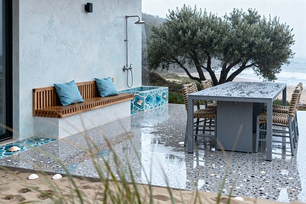 An oceanfront patio shows a polished pebble terrazzo floor with shell inlays, a powder‑coated gray dining table, and four chairs wrapped in thick nautical rope. Against a seafoam stucco wall sits a slatted teak bench with two blue cushions beside a rainfall outdoor shower mounted over a turquoise ceramic mosaic plinth, with rain beading on surfaces and dunes, coastal trees, and the shoreline visible beyond.