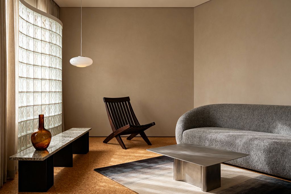 A contemporary living room with taupe walls and cork flooring features a curved glass-block window that filters soft daylight beside sheer beige curtains. A low granite-top bench holds an amber glass vase, while a sculptural dark wood slatted lounge chair faces a rounded gray boucle sofa. A low brushed-steel coffee table rests on a neutral ombré rug, and an opal glass pendant hangs overhead, creating a calm, minimal, textural setting.