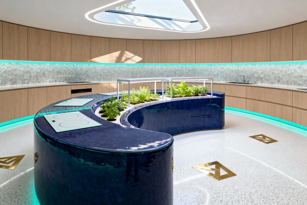 A circular kitchen features light oak cabinetry and a frosted glass mosaic backsplash illuminated by turquoise LED strips. At the center, a glossy cobalt, S-shaped island holds flush white induction cooktops and an integrated herb garden beneath aluminum-framed grow lights. A triangular skylight and recessed ceiling LEDs brighten the terrazzo floor with brass inlays, while a minimalist sink sits along the curved perimeter counter.