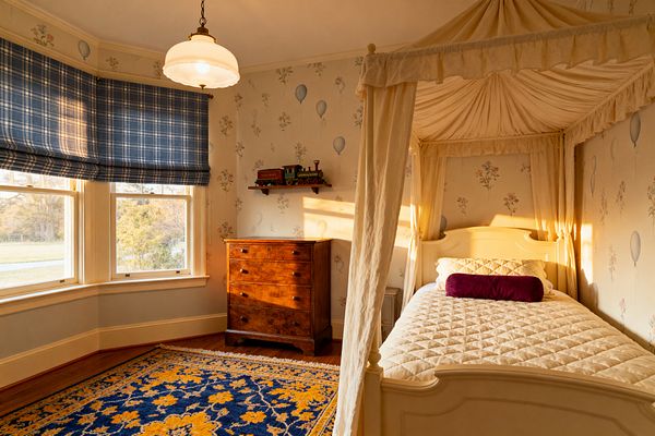 A traditional children’s bedroom features an ivory canopy bed with cream drapery and quilted bedding, accented by a plum bolster pillow. A bay window on the left is dressed with blue plaid Roman shades, allowing warm afternoon light. A cherry wood dresser sits below a shelf with a toy train. The walls have pale floral and balloon wallpaper, a navy-and-gold patterned rug lies on honey wood floors, and a brass milk-glass pendant hangs overhead.