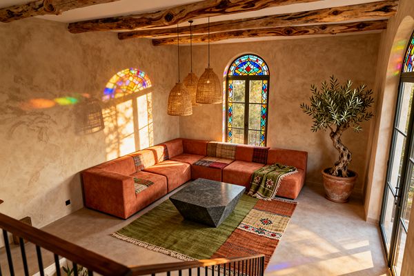 A sunlit living room features an L-shaped terracotta upholstered sofa around a faceted charcoal stone coffee table on a flatweave kilim rug in olive, rust, and cream. Textured plaster walls, exposed raw-wood ceiling beams, and three woven rattan pendants frame arched stained-glass windows that cast colorful light; a potted olive tree and black steel-framed doors complete the relaxed Bohemian scene.