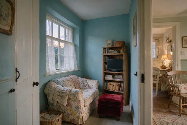 A small cottagecore media room painted in soft blue, featuring a floral slipcovered loveseat under a lace-curtained window. A wooden bookcase with a small television, wicker baskets, and a burgundy velvet ottoman complete the cozy, lived-in space with rustic charm.