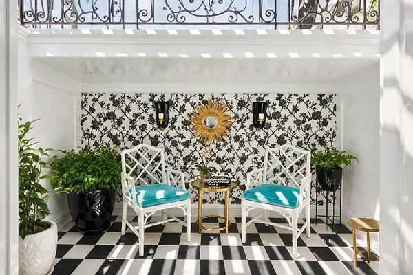 A small outdoor seating nook in Hollywood Regency style with a black-and-white checkered floor, white walls, floral black-and-white wallpaper, and two white lattice-back chairs with bright turquoise cushions. A gold sunburst mirror hangs between black wall sconces, with potted green plants and small brass side tables adding contrast and elegance.