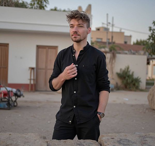 A young man stands confidently outdoors, dressed in a black shirt and pants, with a casual yet stylish demeanor.