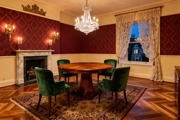 A Victorian-style dining room shows a round mahogany pedestal table surrounded by four emerald-green velvet armchairs on a floral Oriental rug over herringbone parquet. A crystal chandelier hangs above, while a white marble fireplace with brass candle sconces and a gilded crest anchors burgundy damask walls; cream wainscoting, crown molding, and an arched window dressed in lace complete the warm, candlelit setting.
