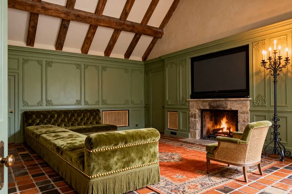 A French Country media room with sage-green paneled walls and carved boiserie features a large flat-screen TV above a rough stone fireplace. An olive velvet, tufted L-shaped chaise with fringe and brass nailheads faces the hearth beside a caned armchair; terracotta floor tiles are layered with a burnt-orange patterned rug, a tall black wrought-iron candelabra stands to the right, and exposed wood beams span a plastered vaulted ceiling, all lit by the fire’s warm glow.