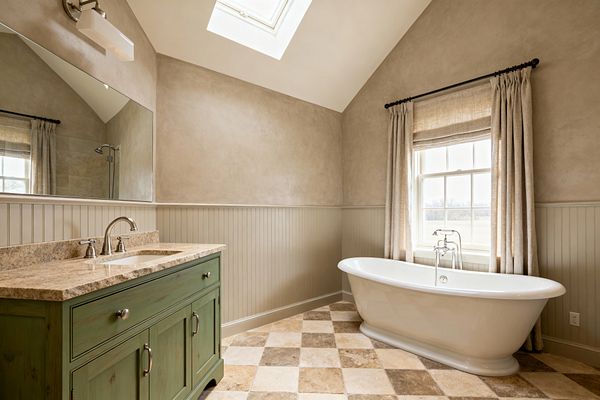 A light-filled transitional bathroom features a freestanding white tub beside a window with linen-look curtains, beadboard wainscoting painted greige, and taupe plaster-look upper walls under a vaulted ceiling with a skylight. On the left, a sage green vanity with a speckled beige granite countertop and brushed-nickel faucet sits beneath a large frameless mirror and a rectangular sconce, while tan-and-cream travertine tiles in a diagonal checkerboard pattern cover the floor.