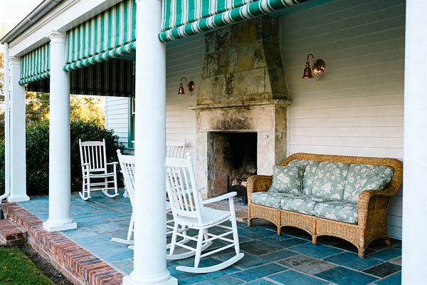 A covered traditional porch features round white columns supporting a roof shaded by green-and-white striped awnings. A large, weathered stone fireplace anchors the space, flanked by copper wall sconces. Two white wooden rocking chairs sit on slate flagstone flooring near a wicker sofa with sage green floral cushions. White clapboard siding, a brick step, and neatly trimmed shrubs frame the patio, creating a calm, classic setting for relaxed outdoor living.