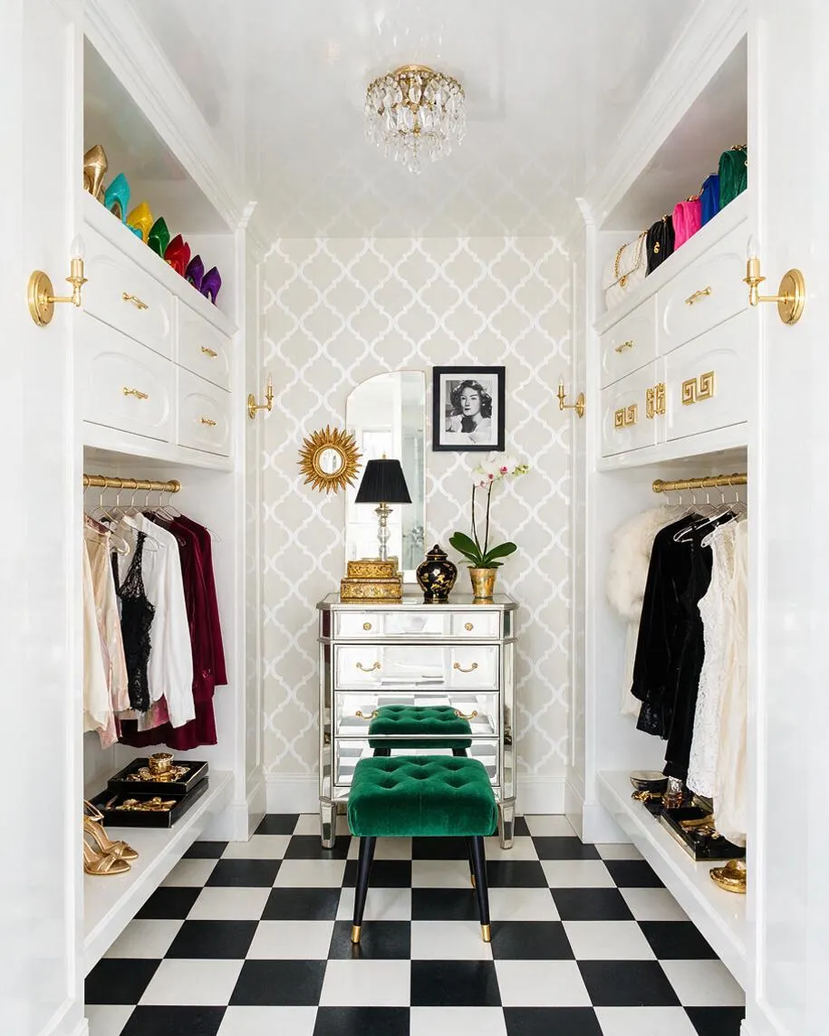 A narrow Hollywood Regency closet with white built-in shelving and drawers, gold hardware, and a black-and-white checkered floor. A mirrored chest, green velvet stool, crystal chandelier, and colorful accessories add a luxe, polished look.