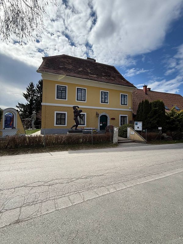 Entrance of the Arnie Museum with a large statue of Arnold Schwarzenegger as a bodybuilder in front of a rustic building.