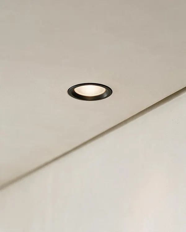 A close-up view of a smooth off-white ceiling with a small recessed downlight featuring a dark trim ring and soft warm illumination. A diagonal wall or beam line crosses the lower half of the image, emphasizing the clean, minimal architectural detail.