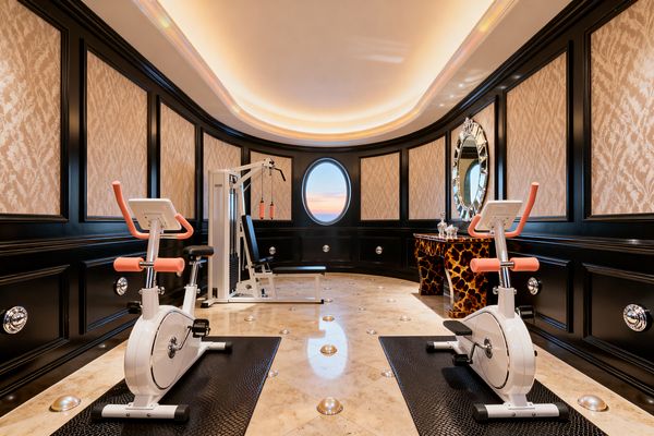 An oval-ceilinged home gym with glossy black paneled walls and champagne damask insets features two white stationary bikes with coral handlebars facing a white weight machine. A polished ivory marble floor with metal inlays sits beneath black textured mats, while cove lighting and a round porthole window create a warm glow. At the far end, a tortoiseshell lacquer console topped with crystal decanters sits below a faceted mirror, adding Hollywood Regency glamour.