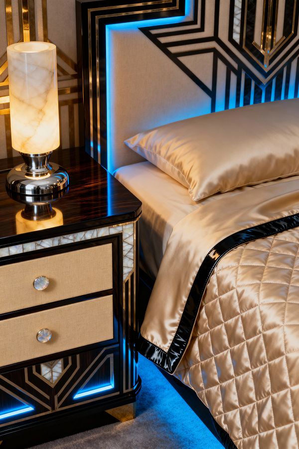 A close-up of an Art Deco bedroom shows a champagne satin quilt with black edging on a bed beside a dark high-gloss nightstand. The nightstand has cream fabric-front drawers with round mother-of-pearl knobs and white onyx geometric inlays. A cylindrical alabaster lamp with a chrome base sits on top. The upholstered headboard displays bold black lacquer and brass linear patterns, backlit by vivid blue LED lighting over a soft gray carpet.