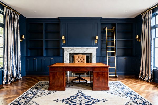 A traditional home office with deep navy paneled walls and built-in bookcases frames a white marble fireplace. A polished mahogany desk with a tan leather writing surface sits centered on an ivory rug with a navy medallion. A camel tufted leather chair faces the room, flanked by four brass sconces. A brass-and-wood rolling library ladder rests to the right. Chevron oak flooring and tall striped navy-and-silver drapes complete the symmetrical, formal space.