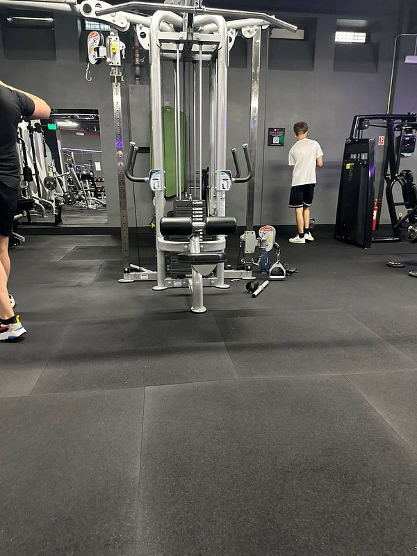 The image captures a gym interior with workout equipment and two individuals engaged in exercise.