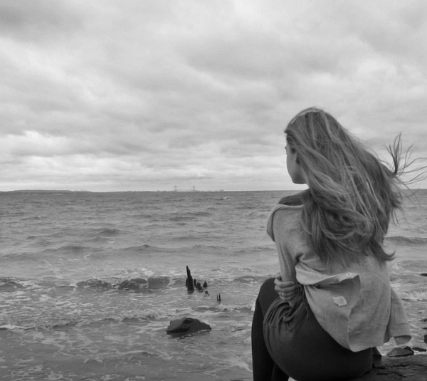 A young woman sits on a rocky shore, gazing out at a turbulent sea under a cloudy sky.
