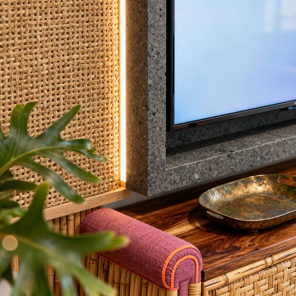 Close-up of a tropical media room detail: a flat-screen TV sits within a speckled gray stone surround. A vertical strip of warm LED light glows between the stone and a woven rattan cane panel. Below, a richly grained wooden top rests on a bamboo-clad cabinet holding a shallow hammered brass tray. A magenta fabric bolster with orange piping sits at the edge, and a leafy plant is visible in the foreground.