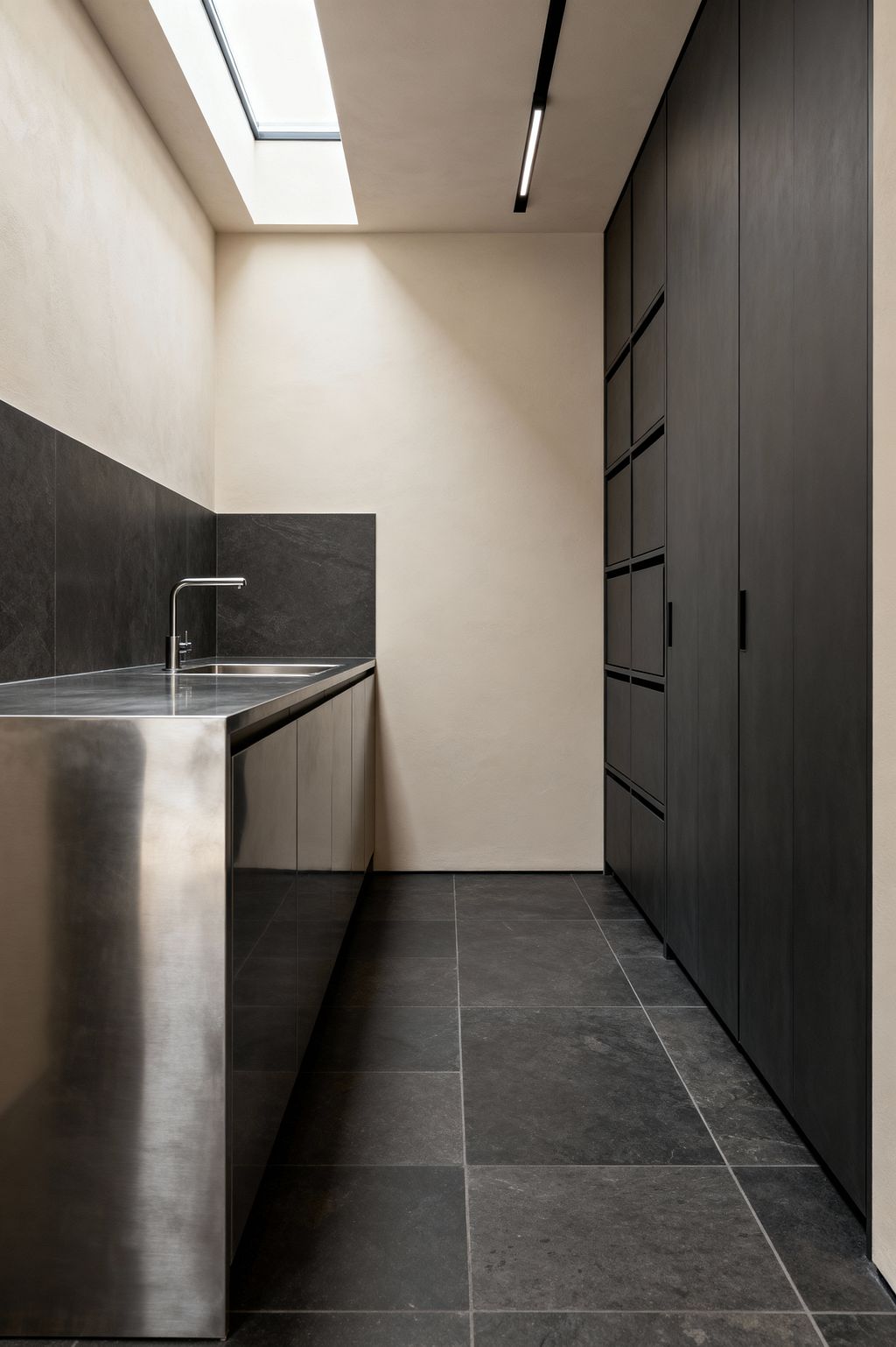 A narrow minimalist laundry features a stainless-steel countertop with an integrated sink along the left wall, backed by a charcoal stone slab. On the right, full-height matte black cabinets with discreet pulls provide storage. Large dark stone floor tiles continue the tone, while warm off-white plaster walls, a ceiling skylight, and a recessed linear light create even illumination and a calm, functional atmosphere.