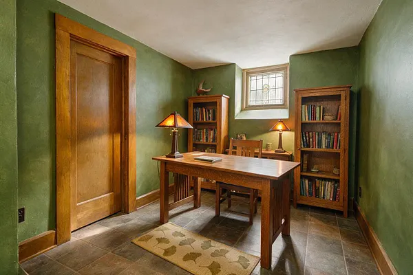 A small basement study in Craftsman style with olive green walls, a wood door, and a tiled floor. The room includes a simple wooden desk, two bookshelves filled with books, warm amber table lamps, a stained-glass window, and a leaf-patterned rug.
