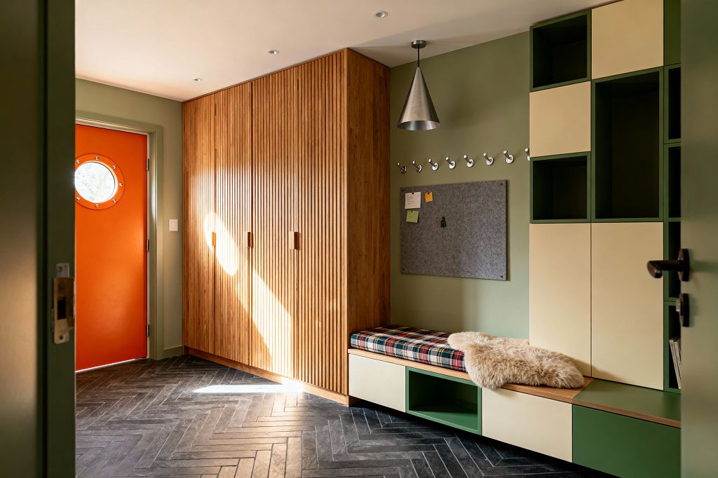 A mid-century modern mudroom with sage green walls and a bright orange door featuring a round porthole window. Tall fluted oak wardrobes and a long built-in bench with cream-and-green cubbies sit over a charcoal herringbone tile floor; above the bench are chrome hooks and a gray felt pinboard, while a conical brushed metal pendant, plaid seat cushion, and sheepskin throw add warmth and practical character.