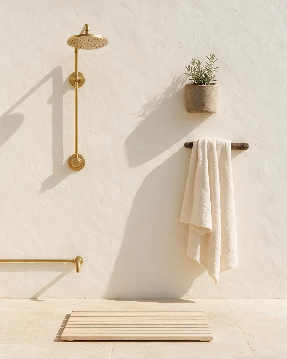 A minimalist outdoor shower area with a brass rain showerhead, matching brass plumbing, a textured white stucco wall, a small wall-mounted planter, and a hanging white towel on a dark wooden peg. A pale slatted wooden mat sits on the light stone floor, creating a calm coastal spa feel.