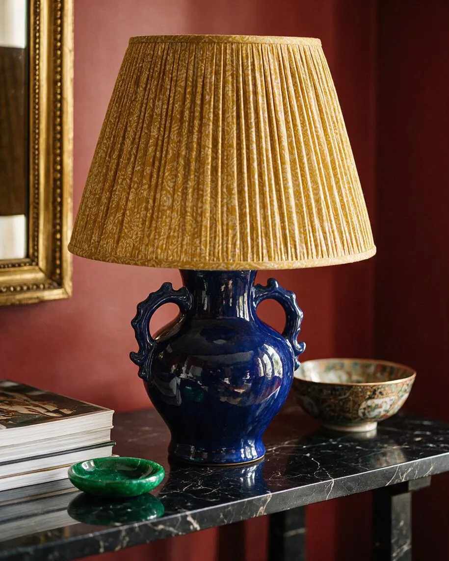 A decorative table lamp with a glossy cobalt-blue ceramic base shaped like a vase with side handles and a pleated gold lampshade. It sits on a black marble console alongside a stack of books, a green dish, and a patterned bowl against a deep red wall.