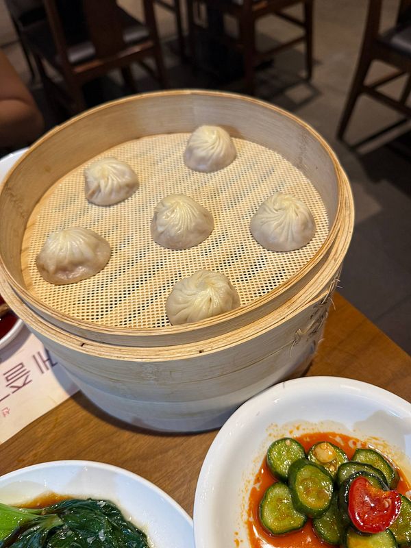 Steamed dumplings served in a bamboo basket with side dishes of greens and spicy cucumbers on a wooden table in a restaurant setting.