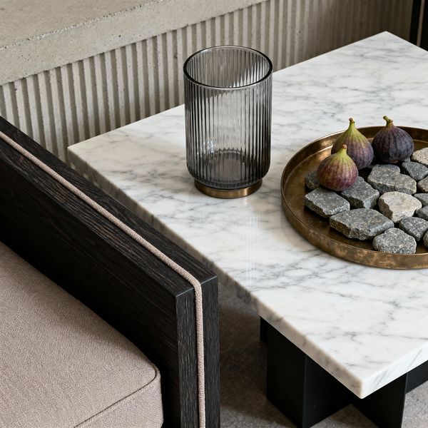 A close-up patio vignette features a rectangular white marble coffee table on a black base. On the surface, a ribbed smoked-glass vase with a bronze foot sits beside a round brass tray holding gray stones and three figs. To the left, an outdoor sofa with an ebonized wood frame and taupe cushions shows tailored piping along the arm. Behind, a light gray fluted concrete bench forms the backdrop, blending natural textures with simple, contemporary lines.