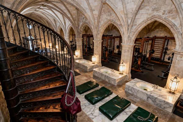 A home gym set within a Gothic stone hall featuring ribbed vaults and pointed arches. A dark wood spiral staircase with a wrought-iron balustrade curves at the left. Behind stone columns, wooden power racks with barbells and dumbbells sit on black rubber flooring. In front, three rectangular white-gray marble basins are paired with brass lanterns, and deep green mats lie on marble platforms. Warm lighting emphasizes the textured limestone walls and ceiling.