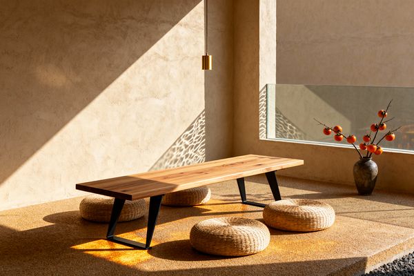 A minimalist Japanese-style dining area with a low rectangular wood table on black metal legs, surrounded by four round woven floor cushions. Sunlight enters through a low window, creating water-like reflections on textured beige plaster walls and a sandy floor. A small brass pendant hangs above the table, and a dark ceramic vase with a branch of orange persimmons sits near the window, beside a narrow edge of dark river stones.