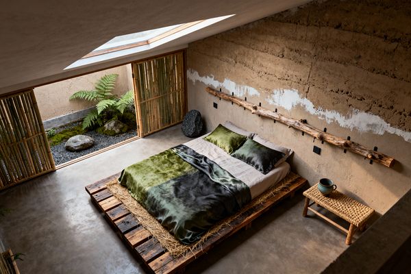 A serene Wabi-Sabi bedroom features a low bed on a reclaimed wood pallet platform layered with straw, dressed in linen sheets and green-to-charcoal satin bedding. Rough earthen plaster walls, a branch peg rail, a woven bench with a ceramic cup, polished concrete flooring, bamboo screens, and a skylit opening to a small gravel-and-moss courtyard with ferns and rocks complete the calm, natural setting.