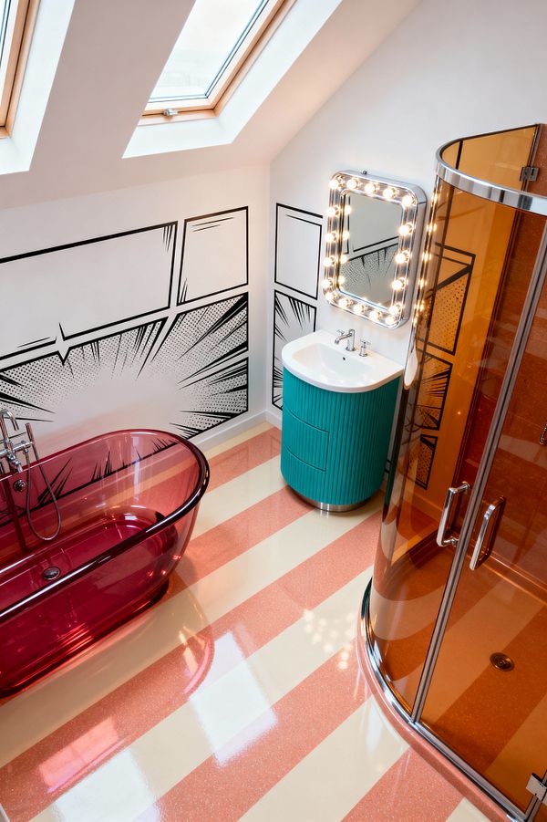 A loft bathroom with a sloped ceiling and skylights features black-and-white comic-strip graphics on white walls, a translucent magenta freestanding acrylic tub with chrome filler, and a corner shower enclosed in amber-tinted glass with chrome framing. A teal fluted vanity with a white basin sits beneath a chrome marquee mirror ringed with bulbs, while a glossy floor in wide peach and cream stripes reflects abundant light for a bold Pop Art look.