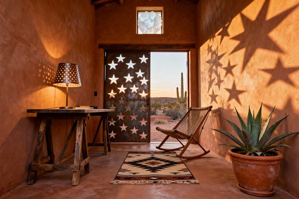 A compact Southwestern home office with terracotta plaster walls features a reclaimed-wood trestle desk topped by a punched-tin lamp and a woven-rope rocking chair. A weathered steel door perforated with stars opens to a desert view with a tall cactus, casting star shadows across the room; a Navajo-style rug lies on the floor and a potted agave sits in a terracotta pot beneath exposed beams and a small clerestory window.