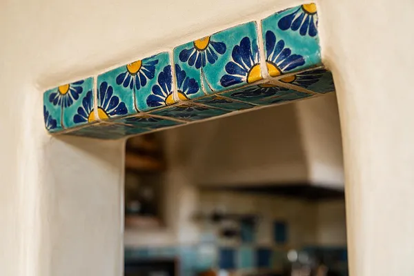A close-up of a doorway or window opening framed with hand-painted turquoise ceramic tiles decorated with navy blue and yellow floral motifs. The surrounding wall is a smooth warm white plaster, and the background beyond the opening is softly blurred.
