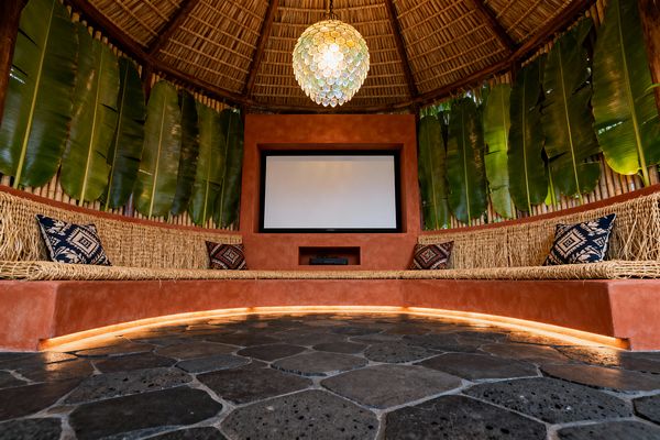 A tropical media room under a round thatched palapa roof features a central projector screen set in a terracotta plaster wall. Curved built-in bench seating with woven seagrass surfaces wraps the space, accented by navy patterned pillows, while vertical banana leaves over bamboo create a lush backdrop, a spherical capiz shell pendant casts warm light, under-bench LED strips glow, and a charcoal basalt flagstone floor anchors the room.
