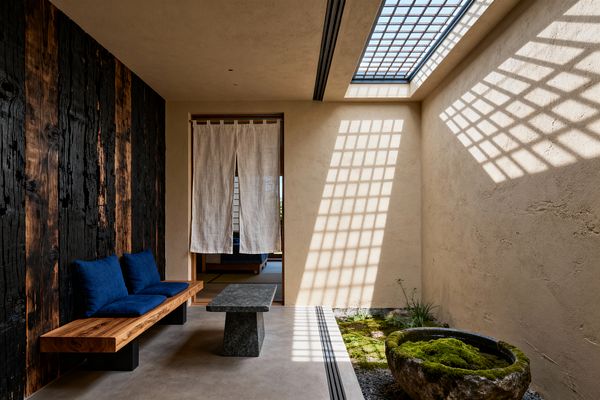 Japanese-style living room featuring a charred cedar (shou sugi ban) wall and a floating solid-wood bench with deep indigo cushions beside a low dark stone table on smooth concrete. Sunlight from a gridded skylight casts lattice shadows across warm earthen-plaster walls, while a small indoor moss garden with a weathered stone water basin sits to the right and a linen noren opens to a tatami room beyond.
