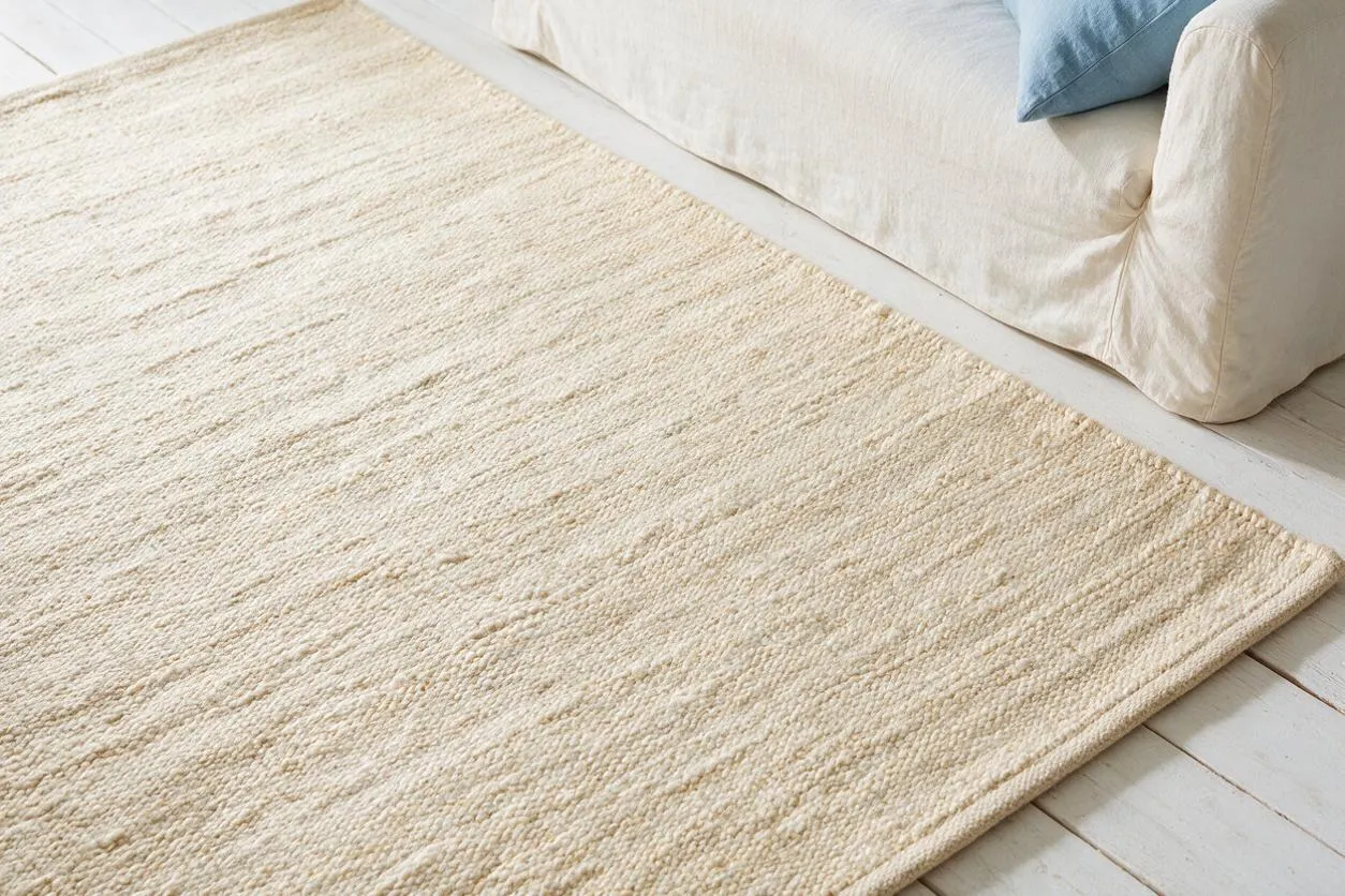 A light cream textured area rug lies on white painted floorboards beside a soft ivory sofa with a pale blue cushion. The rug has a woven, slightly ribbed natural fiber look that adds warmth and a relaxed coastal feel.