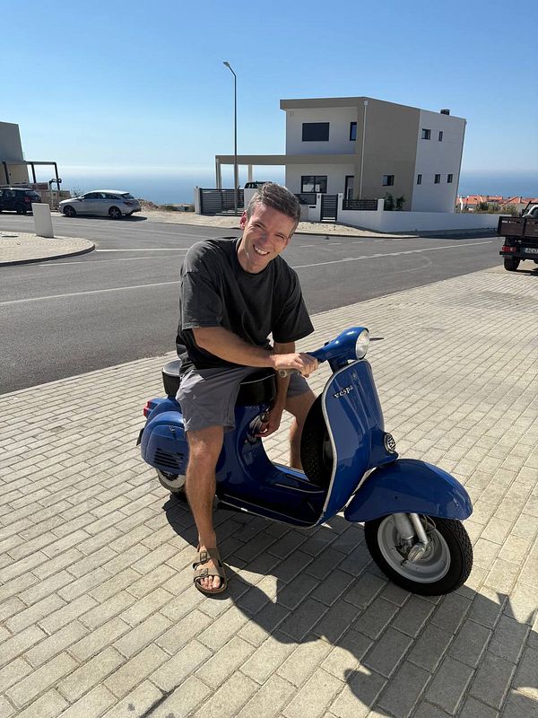 A man is sitting on a blue scooter in a sunny outdoor setting.